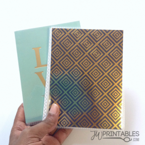 A hand holds two notebooks, the front one with a gold and dark blue geometric square pattern, and the back one a light blue color with gold letters "LOV" visible.