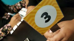 A person holds a yellow card with a white circle displaying the number 3, while another person is seen writing in the blurred background.