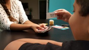 Two people are playing a card game at a table, with one person holding a card that reads "GIVE YOU MY ALL" and the other holding a stack of cards.