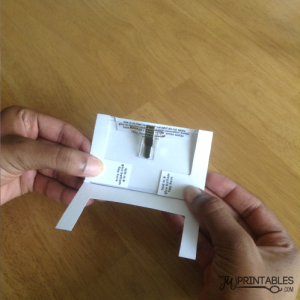 A person's hands hold a white paper craft with a metal clip attached, displaying instructions to "fold in & glue bottom flap here" and other partial text.