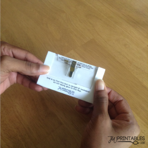 A person's hands hold a white card with a metal badge clip attached, displaying instructions on both flaps for assembly, including "fold in so that this text is upright & readable! do not apply glue to this flap." and the "JM PRINTABLES.COM" logo.