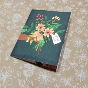 A greeting card with a dark teal background and a floral illustration featuring orange flowers, green leaves, and red berries, with a tag reading 'Get Well Soon!', rests on a light brown surface with white floral patterns.