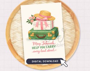 A greeting card featuring an illustration of a straw hat, a white gift box, and a green suitcase, with the text "May Jehovah HELP YOU CARRY every load ahead" and a "DIGITAL DOWNLOAD" button, rests on a wooden tray.