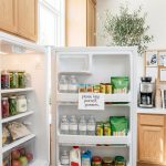 An open refrigerator is filled with bottled water, juice boxes, snack bags, milk, and jars of food, with a note on a shelf that reads "please help yourself, pioneers."