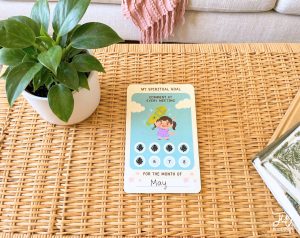 A spiritual goal card for May, featuring a cartoon girl with a microphone and a progress tracker, is displayed on a woven surface with a plant and a book.