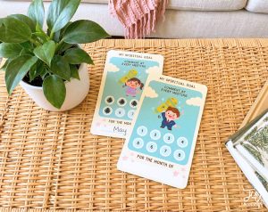 Two spiritual goal tracking cards, one with "May" handwritten, are displayed on a woven rattan surface next to a potted plant, both cards featuring a child with a microphone, the goal "COMMENT AT EVERY MEETING", and numbered circles for tracking.