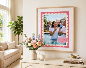 A framed picture featuring a smiling woman in a pool with a pink wavy border and text "SARAH SANTAG ETERNAL HAPPINESS 2026" is displayed on a console table next to flowers, a candle, and books.