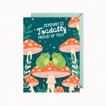 A greeting card features two green frogs sitting on a large red and white spotted mushroom, surrounded by smaller mushrooms, green foliage, and stars, with the text "JEHOVAH IS TOADALLY PROUD OF YOU!".