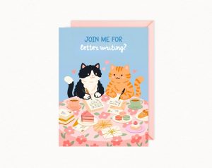 A greeting card features two cartoon cats, one black and white and one orange tabby, sitting at a floral table writing letters, surrounded by tea, cake, macarons, and other pastries, with the text "JOIN ME FOR letter writing?" at the top.