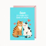 A light blue greeting card features two cartoon cats, an orange tabby and a white cat with black spots, sitting together with small red hearts, and text that reads "love IS A PURR-FECT BOND OF UNION" and "Col. 3:14", with a pink envelope visible behind it.