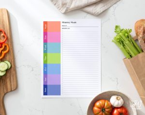 A "Ministry Meals" meal planner with a grocery list is displayed on a white marble counter, surrounded by fresh vegetables like bell peppers, cucumbers, celery, tomatoes, garlic, and a loaf of bread.